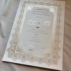 以下が含まれることがあります： 金と銀の装飾が施されたフォーマルな結婚式の招待状。招待状にはアラビア語の筆記体と装飾的なボーダーがあり、「Hajer」と「Mostafa Salman」の名前が大きく表示されています。日付は2026年1月18日、場所はアメリカ合衆国ニューヨークです。