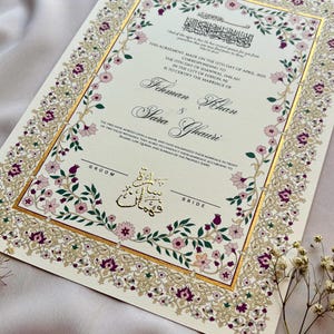 Puede incluir: Invitaci&oacute;n de boda marfil con detalles dorados y borde floral. La invitaci&oacute;n presenta los nombres "Fehman Khan" y "Sara Ghauri" en una escritura elegante, con texto en ingl&eacute;s y &aacute;rabe. El dise&ntilde;o incluye flores rosas y moradas.