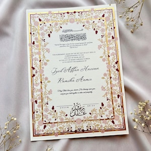 Puede incluir: Una invitación de boda con un diseño floral en rosa y dorado. La invitación incluye texto en inglés y árabe, con los nombres de la pareja, la fecha y el lugar de la ceremonia. La invitación está sobre un fondo blanco.