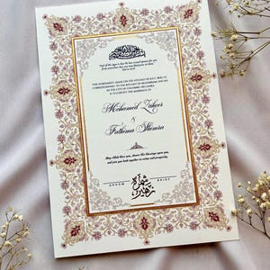 Op de afbeelding: Een trouwkaart met een sierlijke bloemenrand in tinten bordeaux, goud en crème. De uitnodiging bevat elegante kalligrafie met de namen Mohamed Laheer en Fathima Shimra, en bevat tekst in het Engels en Arabisch.