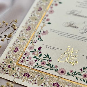 Puede incluir: Invitaci&oacute;n de boda marfil con detalles dorados y borde floral en tonos rosa y morado. La invitaci&oacute;n presenta caligraf&iacute;a elegante y texto en idioma extranjero.