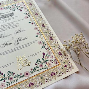 Puede incluir: Certificado de boda de color marfil con detalles en pan de oro y borde floral. El certificado incluye los nombres "Fehman Khan" y "Sara Ghauri", con texto en ingl&eacute;s y &aacute;rabe. Delicadas flores rosas y moradas adornan los bordes.