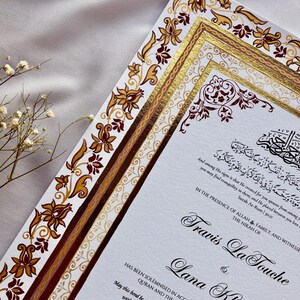 Może przedstawiać: Eleganckie zaproszenie ślubne z ozdobnymi kwiatami i złotymi akcentami. Zaproszenie zawiera kaligrafię z imionami Travis La Touche i Lana Kheder. Projekt zawiera arabski skrypt i dekoracyjne obramowania.