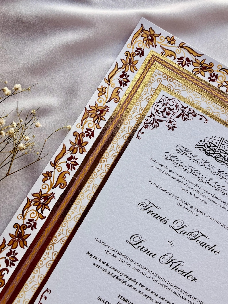 Może przedstawiać: Eleganckie zaproszenie ślubne z ozdobnymi kwiatami i złotymi akcentami. Zaproszenie zawiera kaligrafię z imionami Travis La Touche i Lana Kheder. Projekt zawiera arabski skrypt i dekoracyjne obramowania.