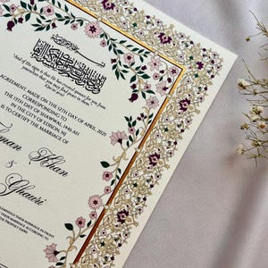 Puede incluir: Certificado de boda de color marfil con un borde floral ornamentado en oro, morado y rosa. El documento incluye caligraf&iacute;a &aacute;rabe y texto en ingl&eacute;s, con los nombres de la pareja y la fecha de la boda.