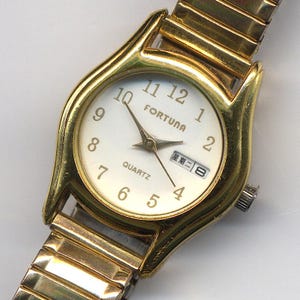 May include: A gold-tone wristwatch with a white face and black hands. The watch has a date window and the words "Fortuna Quartz" printed on the face.