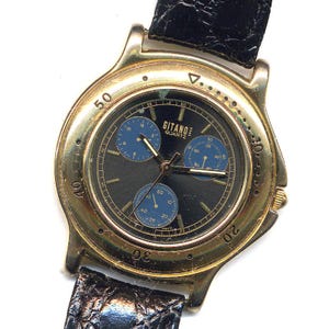 May include: A gold-toned Gitano Quartz wristwatch with a black face and three blue sub-dials. The watch features a black leather band and a gold bezel with minute markers. The watch hands are gold, and the watch face displays the Gitano Quartz logo.