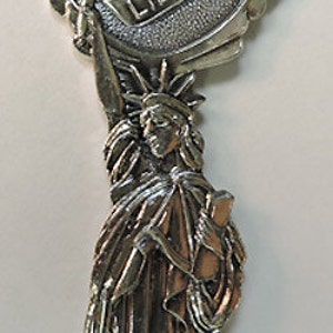 May include: Silver-toned souvenir spoon featuring a detailed depiction of the Statue of Liberty. The handle is shaped like a circle with the words "Statue of Liberty" inscribed on it.