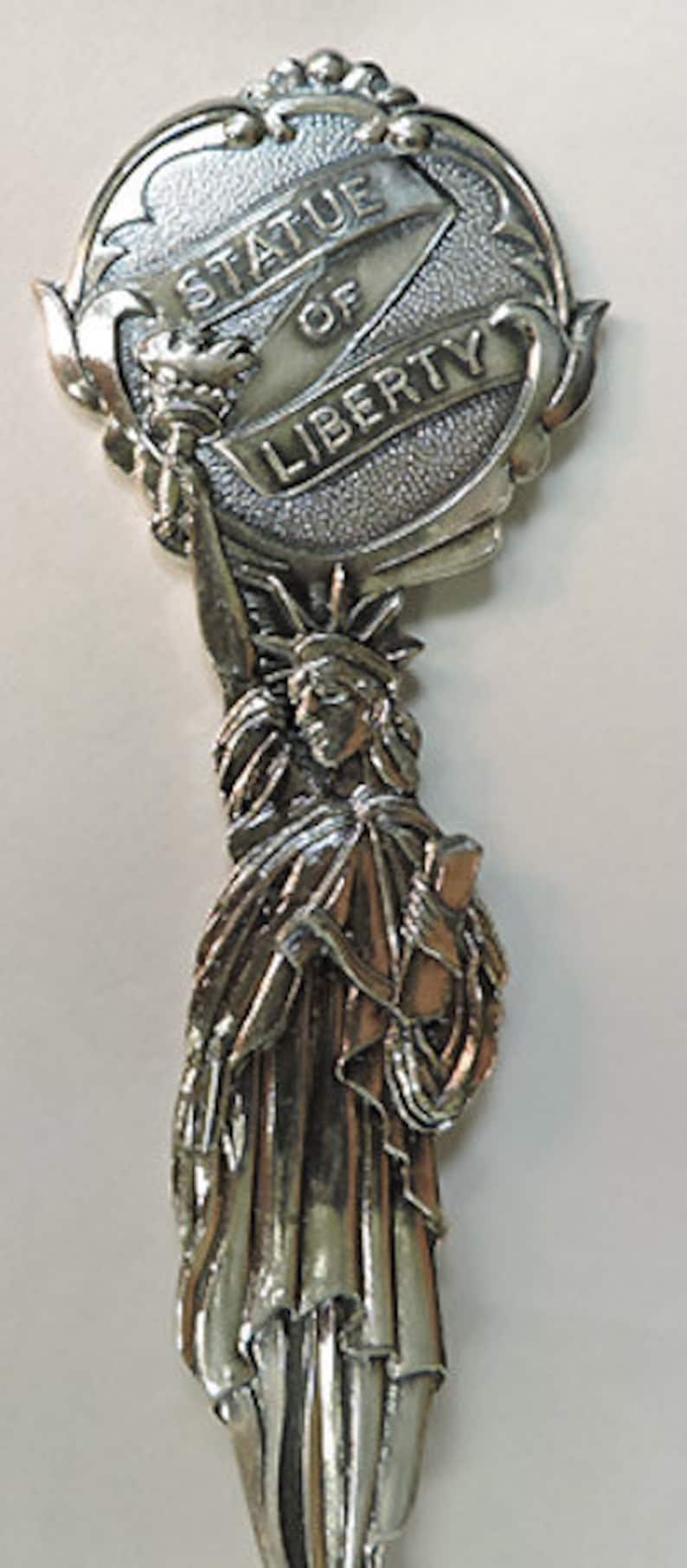May include: Silver-toned souvenir spoon featuring a detailed depiction of the Statue of Liberty. The handle is shaped like a circle with the words "Statue of Liberty" inscribed on it.