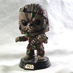 May include: A Funko Pop! figure of Chewbacca from Star Wars, adorned with Christmas lights. The figure is brown with a black base, and the lights are red and green. The Star Wars logo is visible on the base.