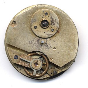May include: Close-up of the inner workings of a vintage pocket watch. The watch face is missing, revealing the intricate gears and mechanisms. The watch is made of brass and has a worn, aged appearance.