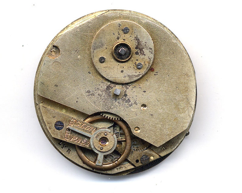 May include: Close-up of the inner workings of a vintage pocket watch. The watch face is missing, revealing the intricate gears and mechanisms. The watch is made of brass and has a worn, aged appearance.