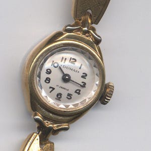 K&ouml;nnte beinhalten: Eine goldfarbene Armbanduhr mit wei&szlig;em Zifferblatt und schwarzen Zeigern. Das Zifferblatt der Uhr hat schwarze r&ouml;mische Ziffern als Stundenmarkierungen und den aufgedruckten Text "DUMAI*" und "17 JEWELS".