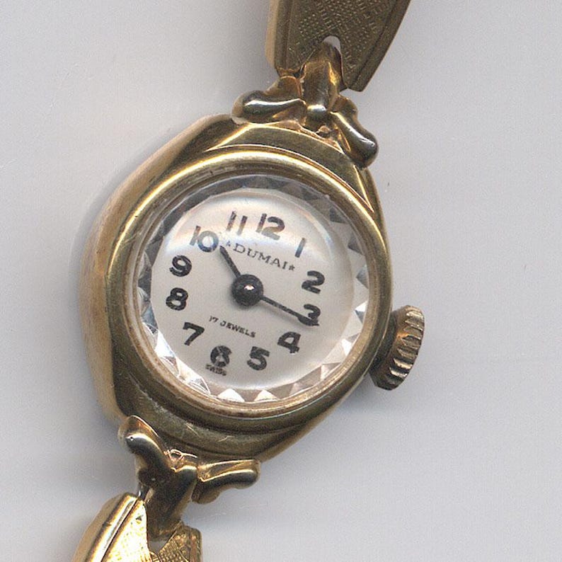 K&ouml;nnte beinhalten: Eine goldfarbene Armbanduhr mit wei&szlig;em Zifferblatt und schwarzen Zeigern. Das Zifferblatt der Uhr hat schwarze r&ouml;mische Ziffern als Stundenmarkierungen und den aufgedruckten Text "DUMAI*" und "17 JEWELS".