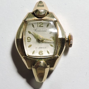 May include: A gold-toned wristwatch with a square face and a white dial. The watch has black Roman numeral hour markers and black hands. The watch face also has the text "Coincert" and "21 Jewels".