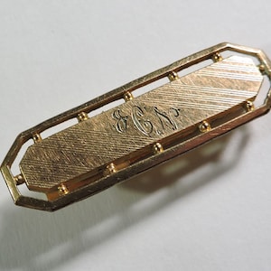 May include: Gold-toned tie bar with a textured surface and a raised border. The tie bar has an engraved monogram "E.C.W."