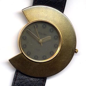 Peut inclure: Une montre-bracelet dorée au design circulaire unique. Le cadran de la montre est de couleur vert foncé avec des marqueurs d'heures noirs et une aiguille des secondes dorée. La montre a un bracelet en cuir noir.