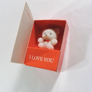 May include: A small white teddy bear with a red bow tie sits inside a red box with the words "I LOVE YOU" printed on the front.
