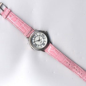 May include: A silver wristwatch with a white face and black numbers. The watch has a pink leather band with a crocodile pattern.