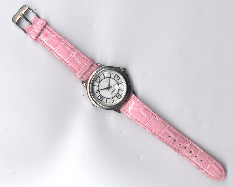 May include: A silver wristwatch with a white face and black numbers. The watch has a pink leather band with a crocodile pattern.