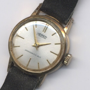 May include: A gold-toned wristwatch with a white face and black leather strap. The watch face has black Roman numeral hour markers, black hands, and the text "KERED DIASHOCK 17 JEWELS".