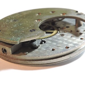 May include: A close-up of a silver and brass clock mechanism with gears and a partially open cover. The mechanism is disassembled and shows the intricate workings of the clock.