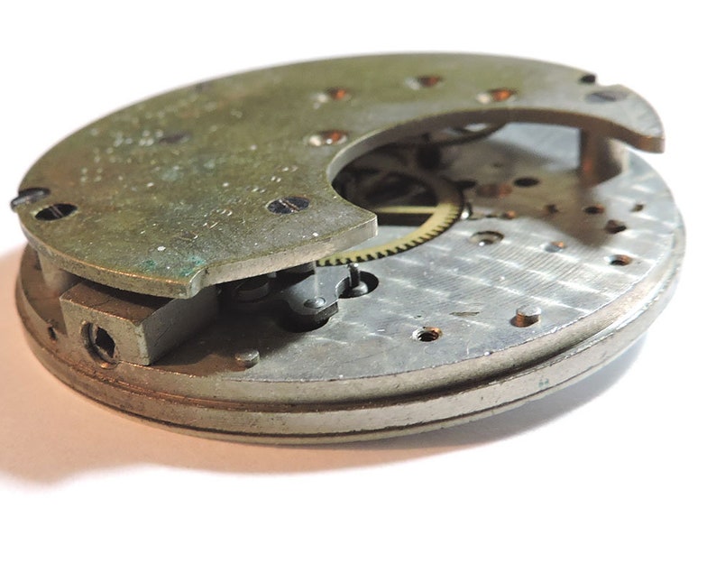 May include: A close-up of a silver and brass clock mechanism with gears and a partially open cover. The mechanism is disassembled and shows the intricate workings of the clock.