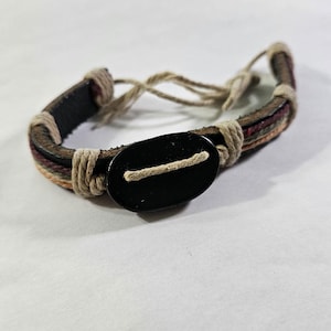 Könnte beinhalten: Mehrstrang-Armband mit schwarzem ovalem Knopfverschluss. Das Armband kombiniert braune, beige und bordeauxfarbene Stränge mit beigen Seildetails. Das Armband ist verstellbar.