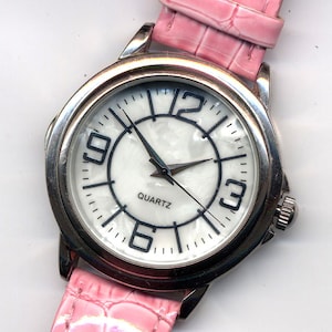 May include: A silver wristwatch with a white face and black numbers. The watch has a pink leather band with a crocodile pattern. The watch face says "QUARTZ".