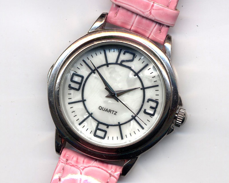 May include: A silver wristwatch with a white face and black numbers. The watch has a pink leather band with a crocodile pattern. The watch face says "QUARTZ".