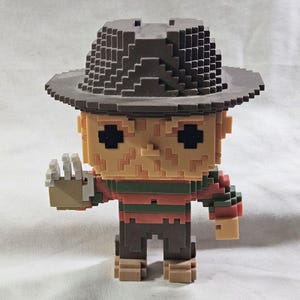 May include: A pixelated Freddy Krueger figure, sporting a brown fedora and a red and green striped sweater. The figure has a tan face and dark eyes, with a hand raised. The design is blocky, resembling a video game character, with a distinctive hat.