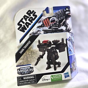 May include: A Star Wars Dark Trooper action figure in its original packaging. The black and red figure is part of the Mission Fleet series. The packaging features the Star Wars logo, Disney+ branding, and a plastic-free packaging label.