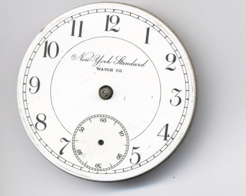 May include: White pocket watch face with black Roman numeral hour markers and black Arabic numeral minute markers. The watch face has the text "New-York Standard WATCH CO."