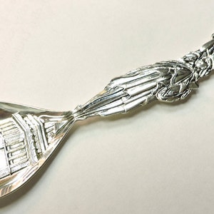 May include: Silver souvenir spoon with a detailed image of the Statue of Liberty on the handle and a depiction of the base of the statue on the bowl. The handle is decorated with a banner that reads "Statue of Liberty".