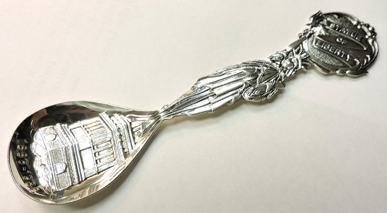 May include: Silver souvenir spoon with a detailed image of the Statue of Liberty on the handle and a depiction of the base of the statue on the bowl. The handle is decorated with a banner that reads "Statue of Liberty".