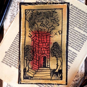 May include: A hand-stitched fabric art piece depicting a red brick tower with a doorway, framed by trees. The artwork uses black line art on a beige background. The text "XD" is visible. The piece is displayed on a background of aged paper.