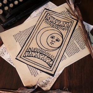 Puede incluir: Una carta de tarot negra y beige con una luna creciente con rostro, rodeada de elementos decorativos. La carta está colocada sobre papel envejecido con texto y una máquina de escribir vintage en el fondo. El tema es místico y vintage.