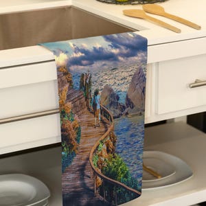 May include: A kitchen towel featuring a scenic coastal image. The design shows a wooden boardwalk winding along a cliffside with a person walking towards the ocean under a cloudy sky. The towel is displayed on a white countertop.