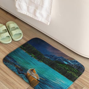 Op de afbeelding: Een rechthoekige badmat met een meerlandschap en een kano. De mat heeft een blauw-groen kleurenpalet, met een bergketen op de achtergrond. Een witte handdoek en lichtgroene sandalen zijn ook zichtbaar.