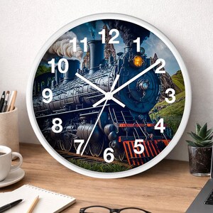 May include: White-framed wall clock featuring a detailed image of a vintage steam train. The clock face displays large white numerals against a backdrop of the train, mountains, and sky. The clock is on a wooden desk with office supplies.