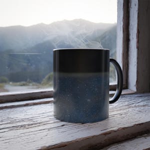 Puede incluir: Una taza de café con degradado negro y azul con motas blancas, emitiendo vapor. La taza está sobre un alféizar de madera pintado de blanco y desgastado, con una cordillera visible a través de la ventana.