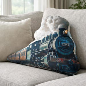 May include: A decorative pillow shaped like a vintage steam train. The pillow features a detailed illustration of a blue and green locomotive with the number 7845 on the front. The train is emitting white smoke, and the pillow is resting on a gray sofa.