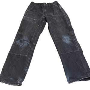 May include: Black work pants featuring a double-knee construction with reinforced stitching and metal rivets. The pants show signs of wear, including a distressed, frayed hole on the left knee and worn fabric at the bottom hems of the legs.