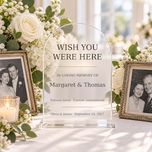 May include: A clear, arched acrylic memorial sign with black text reading WISH YOU WERE HERE, IN LOVING MEMORY OF Margaret & Thomas, Forever loved · Forever remembered, and Olivia & James · September 14, 2027, standing on a rectangular base.