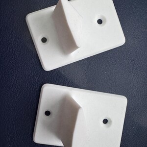 May include: Two white, rectangular wall hooks with a triangular projection. Each hook has two pre-drilled holes for mounting. The hooks are made of a solid, matte white material and are set against a dark blue background.