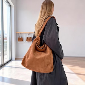 May include: A large, tan suede shoulder bag with a long strap, worn over the shoulder. The bag has a rounded shape and a small pocket on the front. In the background, there are smaller matching bags hanging on a rack.