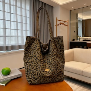 以下が含まれることがあります： 茶色と黒の色合いのヒョウ柄の大きなトートバッグ。バッグには長いハンドルと前面に金色のエンブレムが付いています。バッグは茶色のテーブルの上に置かれています。