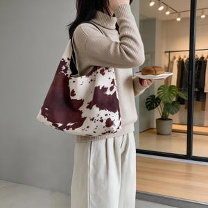 May include: A large tote bag with a cowhide pattern in shades of brown and white. The bag has a black strap and is carried over the shoulder. The bag is made of a sturdy material and is suitable for carrying groceries or other items.