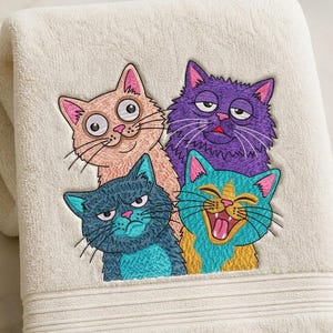 Pode incluir: Toalha de banho creme com um desenho bordado de quatro gatos de desenho animado. Os gatos são de várias cores, incluindo rosa, roxo, azul e amarelo, cada um com uma expressão facial diferente. A toalha tem uma superfície texturizada.
