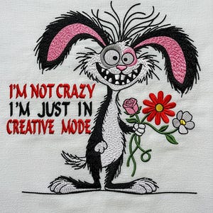 Könnte beinhalten: Gesticktes Design mit einem Cartoon-Kaninchen mit schwarzen und rosa Ohren, das Blumen hält. Das Kaninchen hat ein breites Grinsen und den Text "I'M NOT CRAZY I'M JUST IN CREATIVE MODE" in Rot. Das Design befindet sich auf weißem Hintergrund.